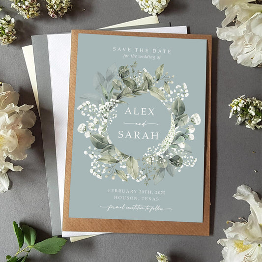 Baby&#39;s breath save the date card with a choice of envelopes featuring an array of gypsophila and eucalyptus greenery in a wreath on a dusty blue background