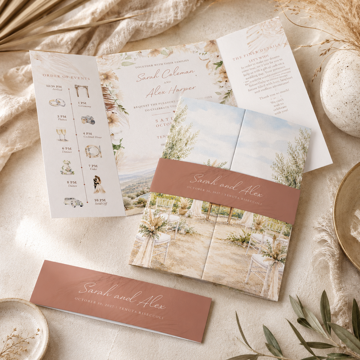 A styled flat-lay of a wedding invitation suite on a cream fabric background with dried botanicals and small ceramic dishes. The set includes a rose-gold and white invitation card, a folded invitation with a wedding venue photo, an itinerary card, and a matching RSVP card.