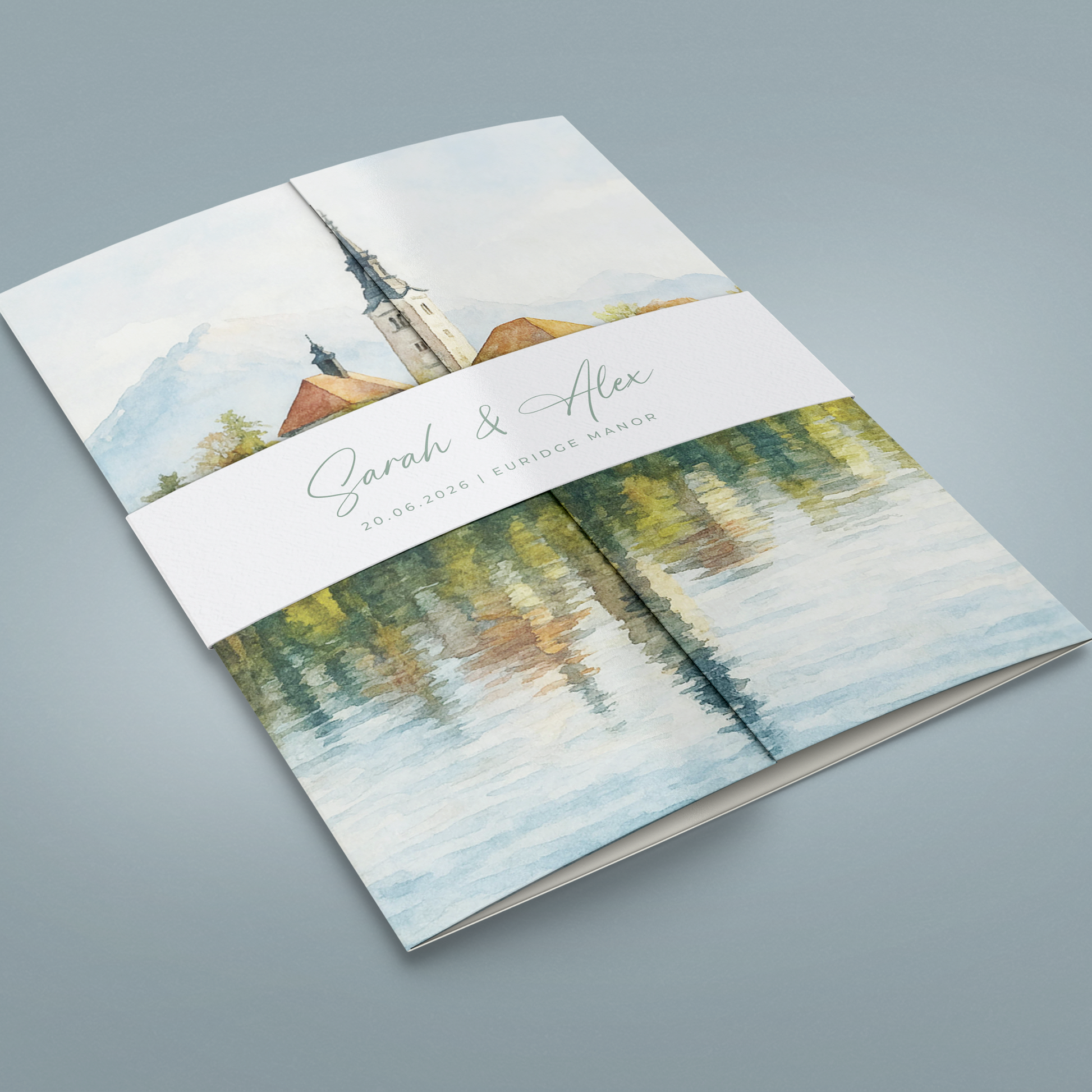 A printed wedding program or booklet cover shown at an angle on a soft gray background. The cover features a watercolor-style illustration of a church tower over reflective water, with a white paper band across the middle reading 'Sarah & Alex' and smaller text below.