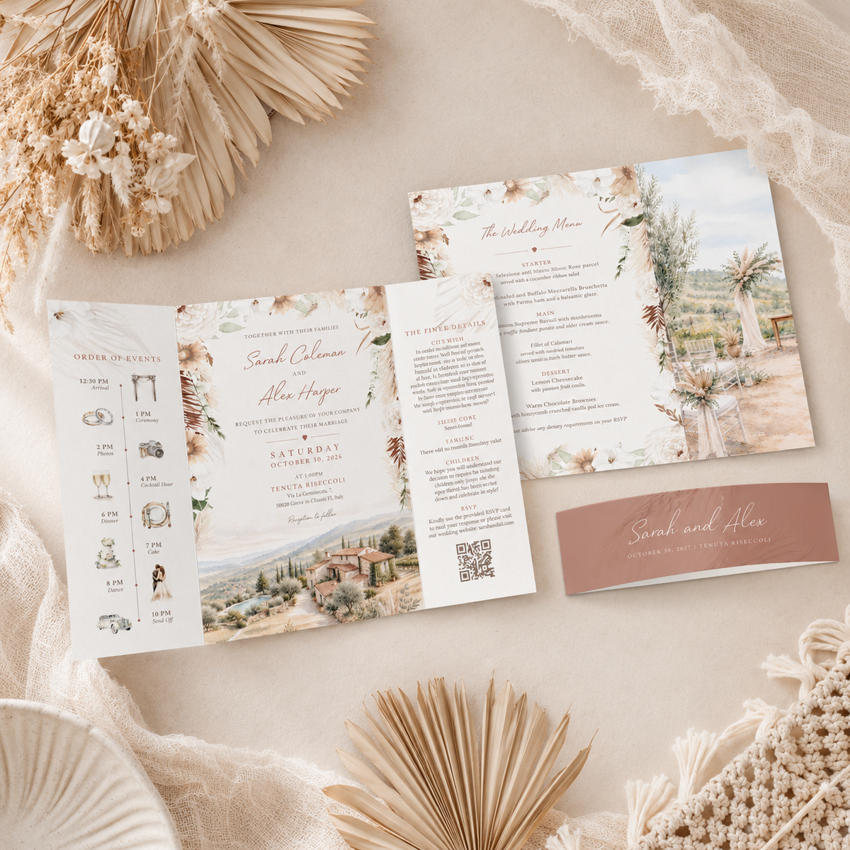 A styled flat-lay of a wedding invitation suite on a beige fabric background with dried flowers and lace. The set includes an open tri-fold invitation with floral and scenic illustrations, a separate folded insert page, a QR code panel, and a dusty rose business card.