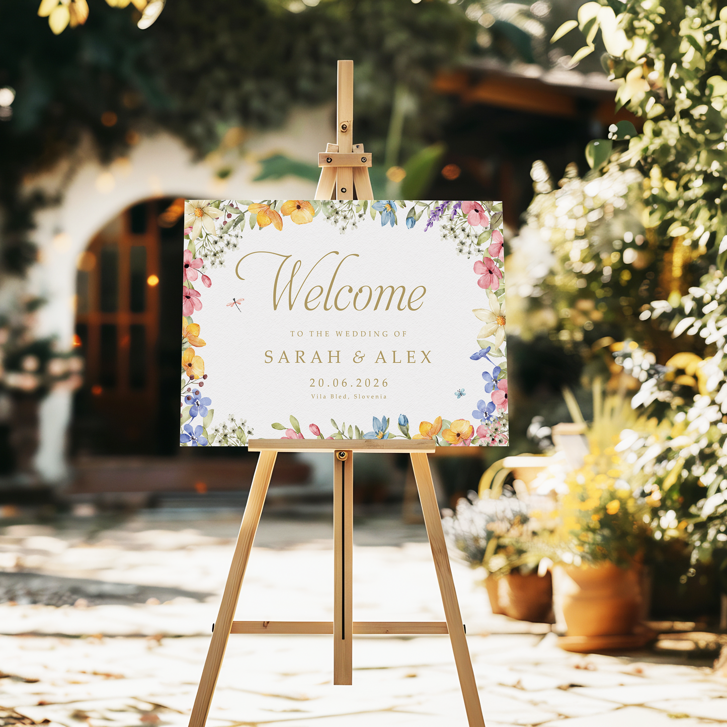 wedding welcome sign with a garden floral border and gold script text reading 'Welcome to the wedding' stands on a wooden easel in an outdoor garden setting.