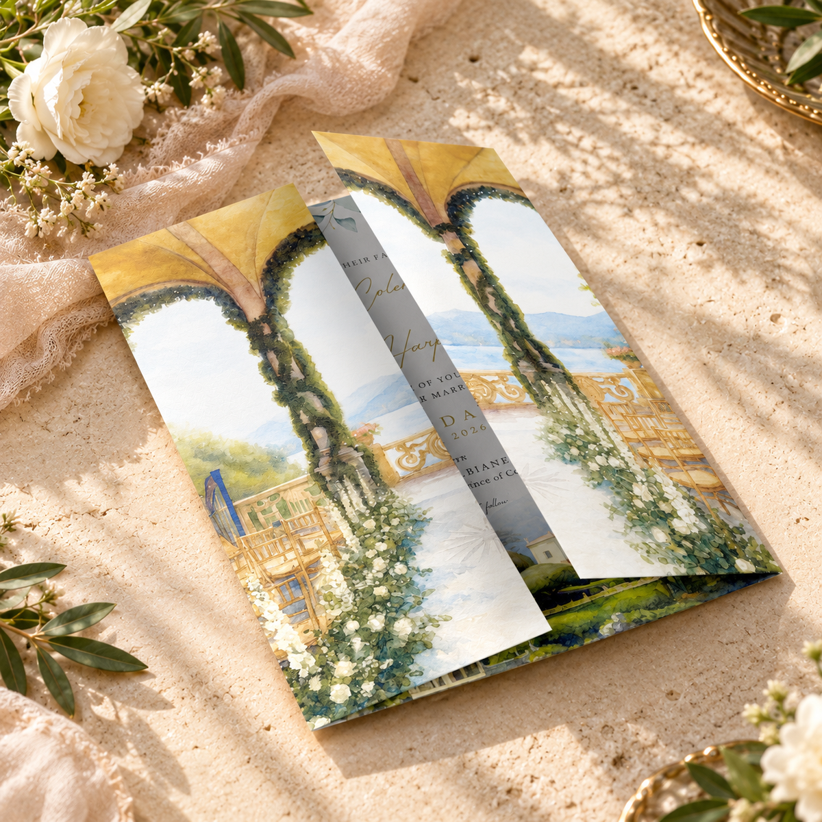 A folded printed invitation card featuring white floral flowers with an arched garden pavilion scene with greenery, white flowers, and a blue sky, styled on a sandy surface with soft fabric and floral props.